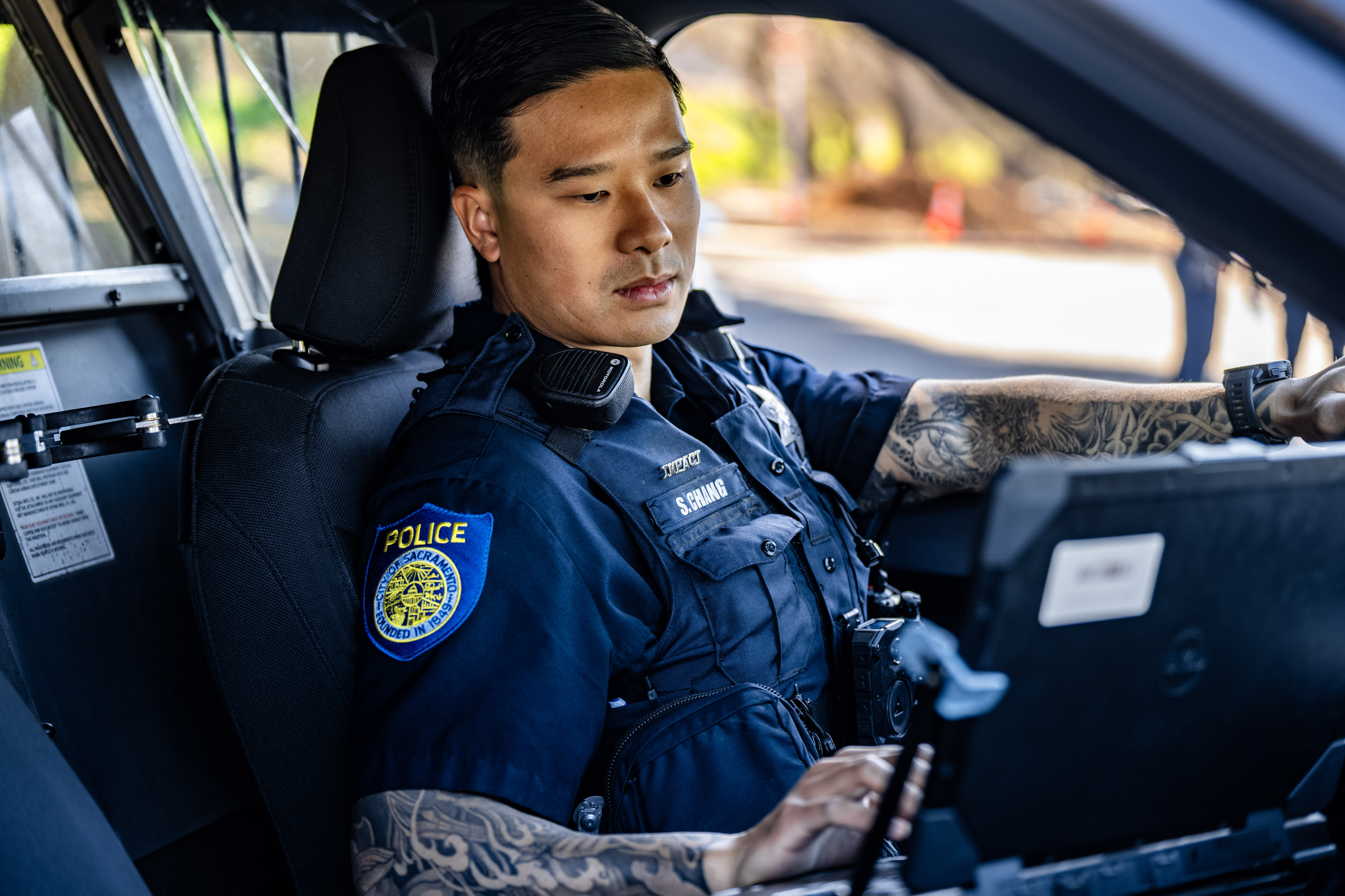 A photo of a Sacramento Police Department officer in a short-sleeved uniform working on their laptop in a patrol car with a visible tattoo "sleeve"